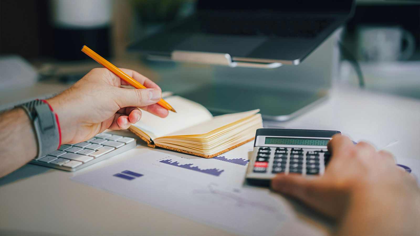Hands using a calculator and a notebook over a desk with financial charts and a laptop in the background